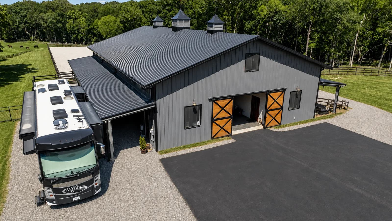 Aerial view of a dark gray engineered steel barn with three cupolas, wood cross-brace sliding doors, a covered RV bay, and horse pasture behind it.
